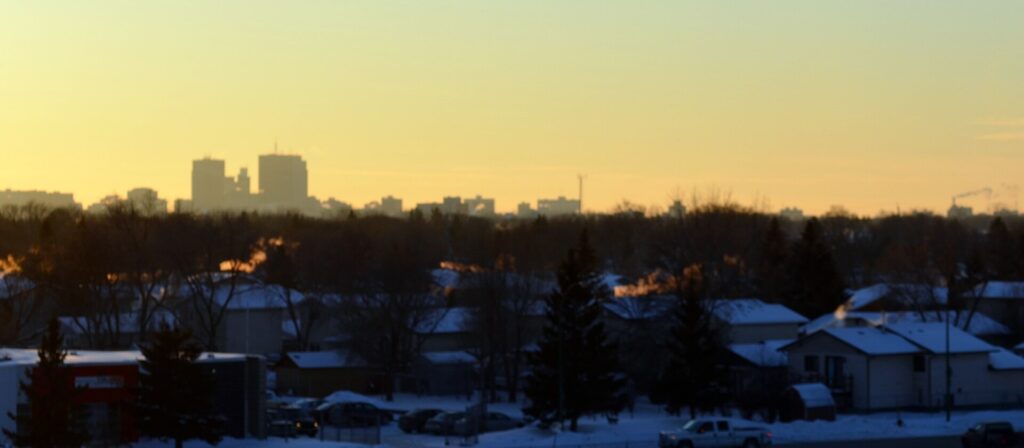 Post-war rooftops in Winnipeg's Kildonan area at sunset, with the downtown skyline in the distance